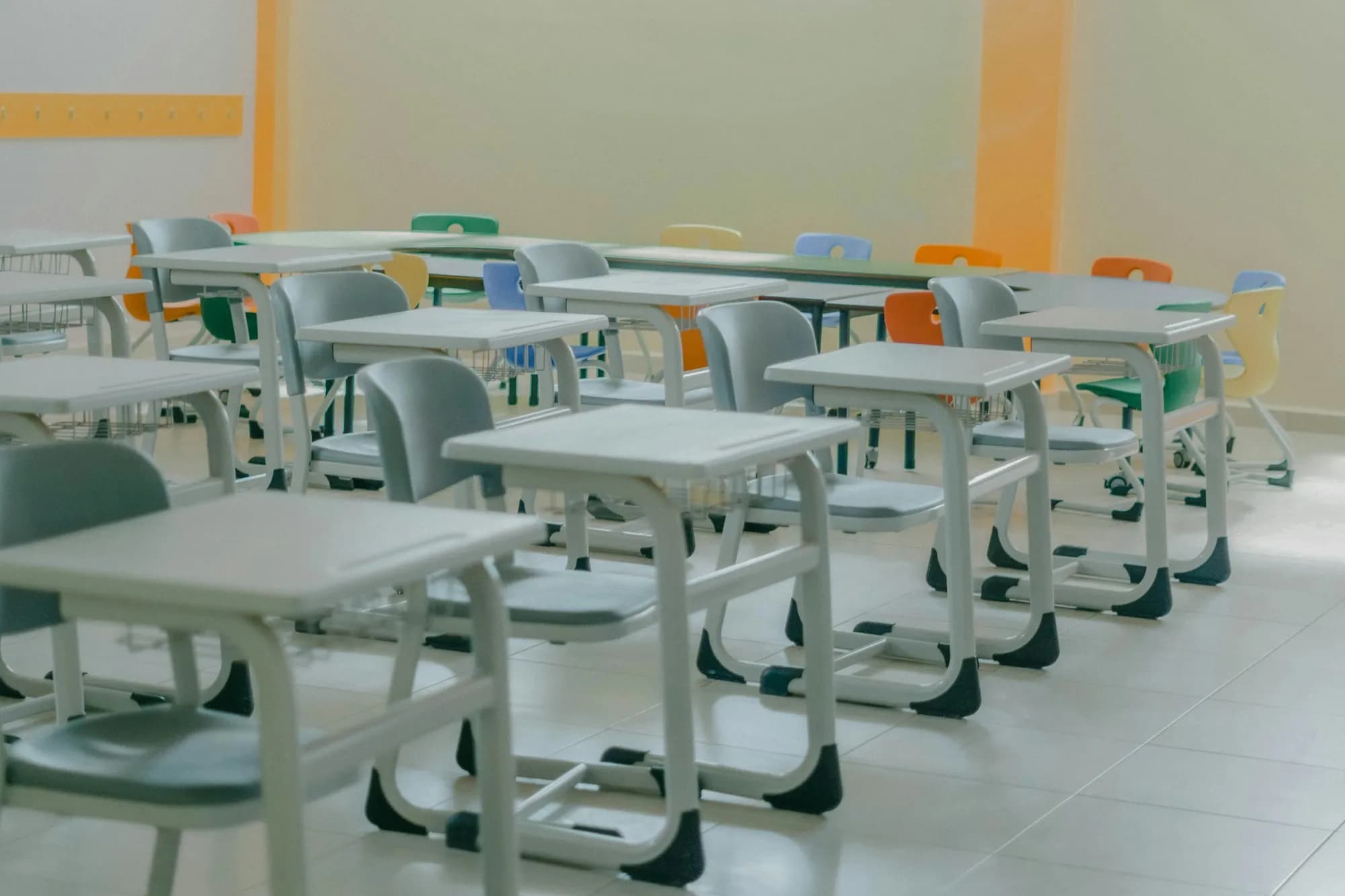 An empty, sunlit classroom with rows of white desks and grey-green chairs