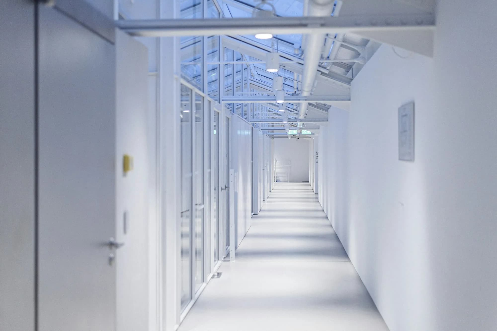 A long white corridor under a glass roof, with doors along both sides